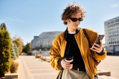 Güneş gözlüklü genç bir adam yoğun bir meydanda kahvesini yudumlarken telefonunu kontrol ediyor..