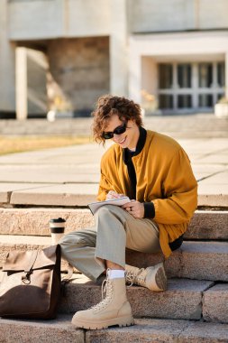 Moda kıyafetli genç bir adam kampüs merdivenlerinde rahatlıyor, defterine çizim yapıyor ve kahvesini yudumluyor..