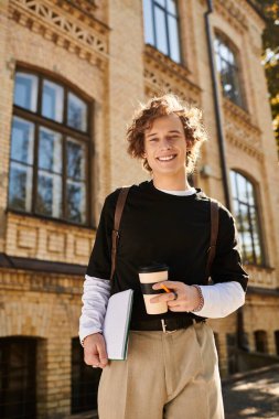 Charming young man holds a coffee cup and notebook while walking past historic campus architecture. clipart