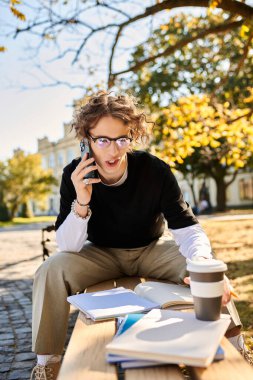 Young man sits outdoors on a sunny autumn day, focused on his phone call and studies. clipart