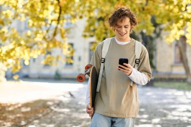 Parkta akıllı telefonuna bakarken sıradan bir şekilde gülümseyen genç bir adam..