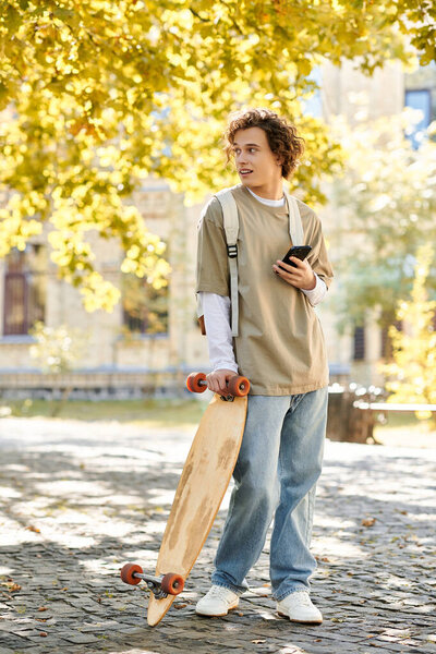 A handsome young man smiles at his phone while holding a skateboard in a sunny, leaf filled park.