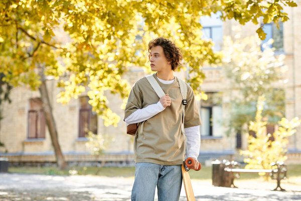 A young man dressed in casual attire stands beneath golden autumn leaves, ready to skate.