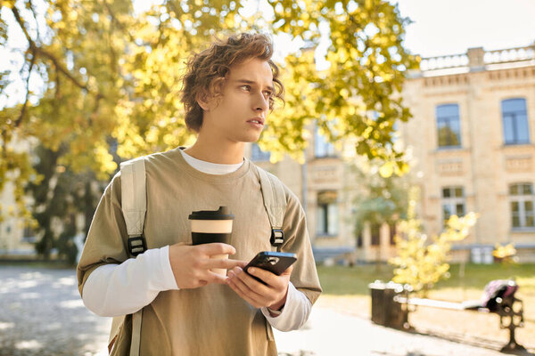 Young man stands outside in a sunny park, sipping coffee and engaged with his smartphone.