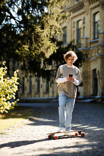 A young man skateboards along a cobblestone path, focused on his smartphone amidst a sunny day.