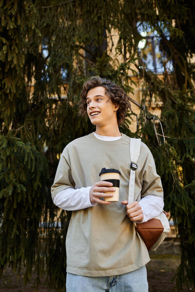 Smiling young man with curly hair holds a coffee cup among lush trees on a sunny day.