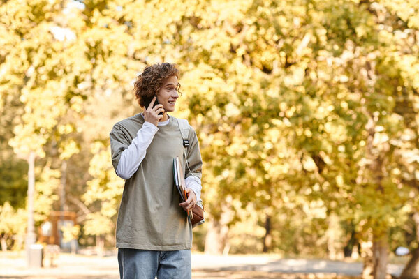 A young man in stylish casual wear chats on his phone while walking through a park.