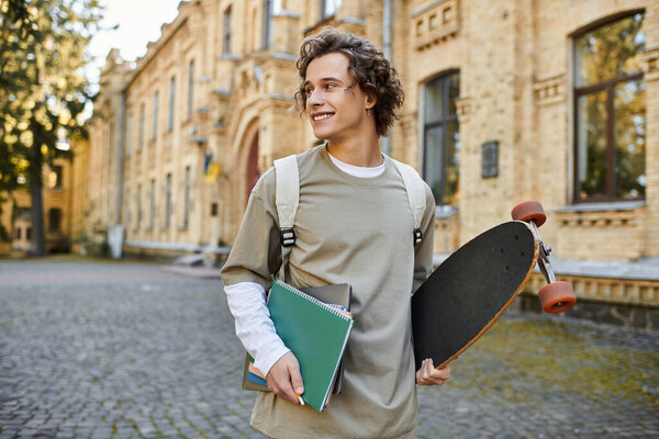 A cheerful young man walks through a classy university courtyard with a skateboard and books.