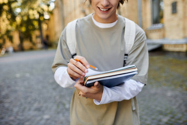 A stylish student smiles as he writes in his notebook outside a historic building on a sunny day.