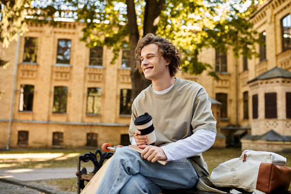 A young man sits on a bench, happily sipping coffee among university buildings.