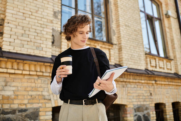 A stylish young man outside a brick building, sipping coffee and reviewing his notes.