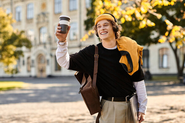 A cheerful young man strolls through a lively, leaf strewn campus with a coffee cup.