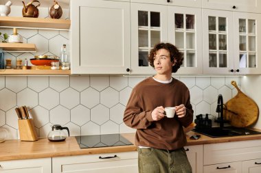 Sıcak, modern bir mutfakta, genç bir adam beyaz bardaktan yudumlayarak öğrenci hayatını yansıtıyor..