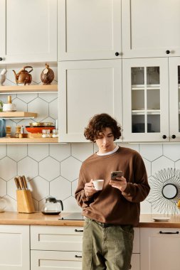 Kahverengi kazaklı genç bir adam evde telefonunu kontrol ederken elinde bir fincan kahve tutuyor..