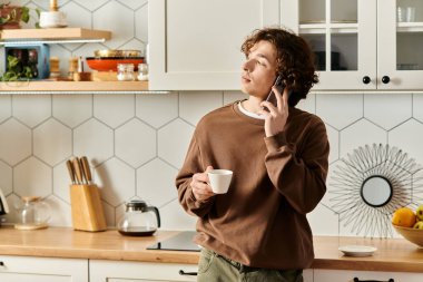 Kahverengi kazaklı genç bir adam telefonda konuşurken elinde bir bardak tutuyor..