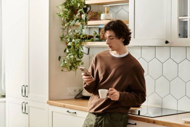 Kıvırcık saçlı öğrenci şık bir mutfakta akıllı telefonuna odaklanırken kahvenin tadını çıkarıyor..