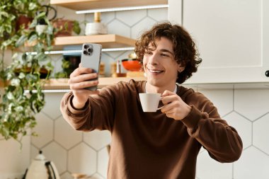 A young man smiles as he takes a selfie, holding a cup in his modern kitchen during daylight. clipart