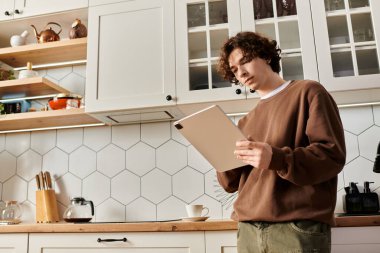 Kahverengi kazaklı genç adam modern mutfak alanında bir tablet üzerinde çalışıyor..