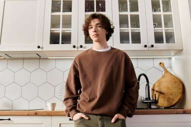 Kahverengi kazaklı genç bir adam şık bir mutfakta duruyor, kendine güven ve sükunet yayıyor..