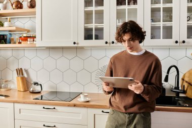 Kahverengi kazaklı genç adam şık mutfağında huzur içinde oturup tabletine odaklanmış..