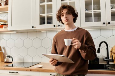 Kıvırcık saçlı öğrenci, şık mutfağında tablet kullanırken kahveyle rahatlıyor..