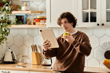 Genç bir adam bir tablet ve bir elma tutuyor, şık bir mutfakta çalışmalarına odaklanmış..