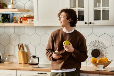 Kahverengi kazaklı genç adam modern mutfağında gevşiyor, telefonunu karıştırıyor..