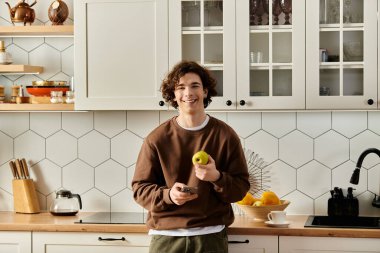 Kahverengi kazaklı genç bir adam şık mutfağında duruyor, elinde bir elma ve telefon var..