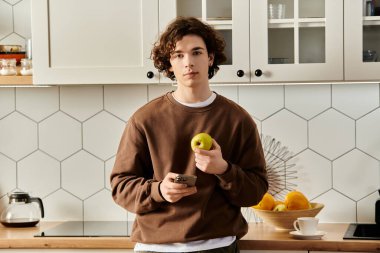Kahverengi tişörtlü genç bir adam, elinde bir elma ve bir telefon tutuyor, rahat dairesini gösteriyor..