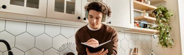 Şık bir mutfakta, kahverengi kazaklı genç bir adam müzik dinlerken notlar yazar.