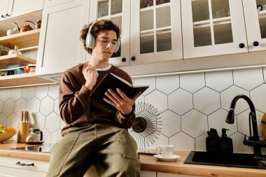 Kahverengi kazaklı odaklanmış genç bir adam şık mutfağında oturmuş kitap okuyor..