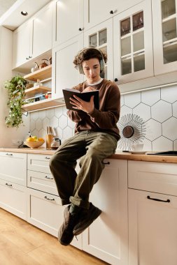 Kahverengi kazaklı genç bir adam modern mutfağında rahatça oturup tabletine odaklanır..