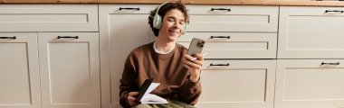 Kahverengi kazaklı genç adam telefonunu kullanarak gülümsüyor ve rahat bir dairede pankartta dinleniyor.