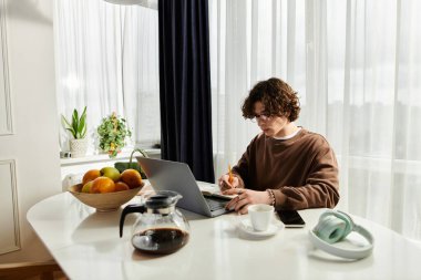 Kıvırcık saçlı genç bir adam yakında taze meyveleri olan gösterişli bir masada okuyor ve yazıyor..