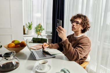 Kahverengi kazaklı genç adam şık bir masada ders çalışırken akıllı telefonuyla etkileşime girer..