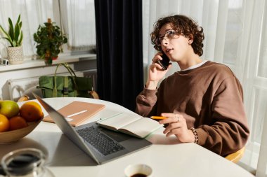 Kahverengi kazaklı genç adam modern apartman masasında çalışırken telefonla konuşuyor..