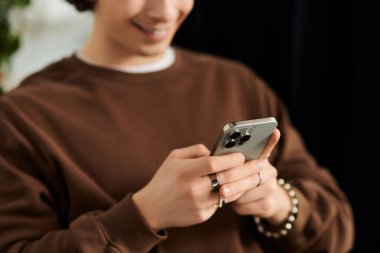 Kıvırcık saçlı genç bir adam telefonunda okuyor, rahat bir şekilde kahverengi bir kazak giyiyor..