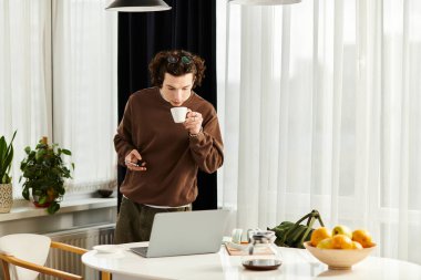 Kahverengi kazaklı genç adam evde dizüstü bilgisayarına odaklanırken kahvesini yudumluyor..