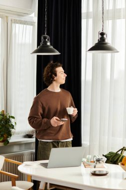 Kahverengi kazaklı genç bir adam evinin penceresinin yanında kahvenin tadını çıkarıyor..