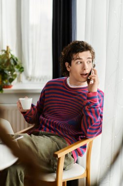 İkili olmayan genç bir insan, parlak bir dairede kahve yudumlarken telefonla konuşuyor..