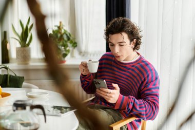 Kıvırcık saçlı, çift kişilikli olmayan kişi, canlı bir dairede telefonlarını kontrol ederken kahvesini yudumluyor..