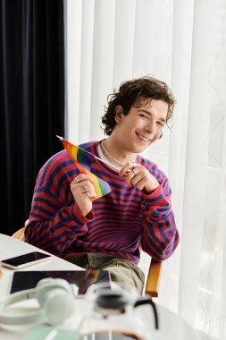 İkili olmayan genç bir insan şık bir dairede gökkuşağı bayrağı tutarken gülümsüyor..