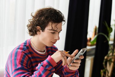 Çizgili tişörtlü genç, ikili olmayan kişi şık bir dairede telefonuna odaklanıyor..