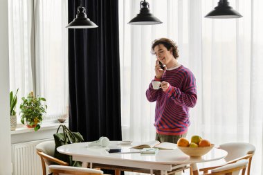 İkili olmayan genç bir insan, modern evinde çayını yudumlarken telefonla konuşuyor..