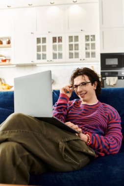 Renkli çizgili tişörtlü, ikili olmayan genç bir insan evde rahatlıyor ve dizüstü bilgisayarına odaklanıyor..