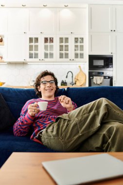 İkili olmayan genç bir birey kanepede rahatlar, kahvesini yudumlar ve sıcak bir anı kucaklar..