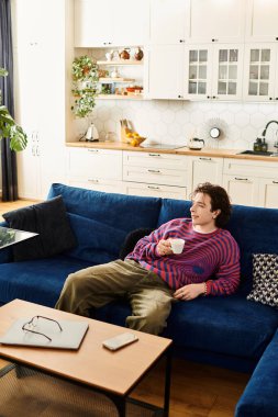 İkili olmayan genç bir insan koltukta rahatlar, şık bir dairede kahvesini yudumlar..