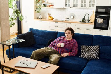 Canlı çizgili bir eşofman üstü içinde, genç bir ikili olmayan kişi evde uzanırken kahvenin tadını çıkarıyor..