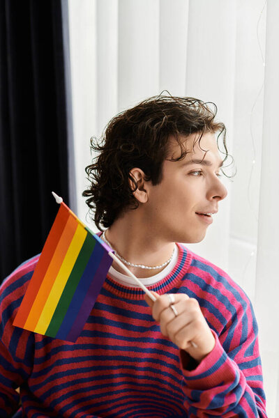 Young non-binary individual with curly hair holds a rainbow flag while sitting in a modern space.