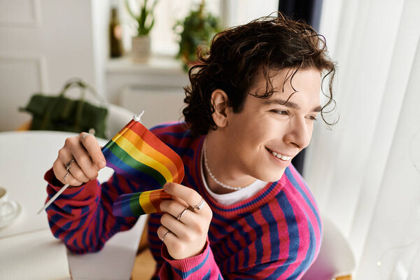 A young non binary individual with curly hair smiles while holding a rainbow flag in a cozy space.